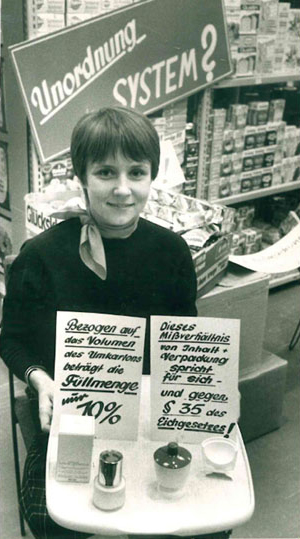 Eine Frau zeigt auf einem Foto in Graustufen diverse Mogelpackungen in den 70-er Jahren.