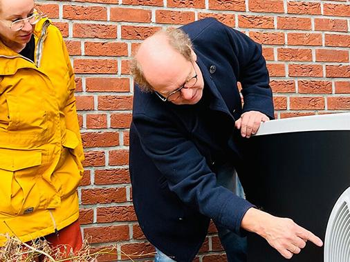 Energieberater zeigt gebeugt auf eine Wärmepumpe. Links daneben steht eine Frau und schaut zu.