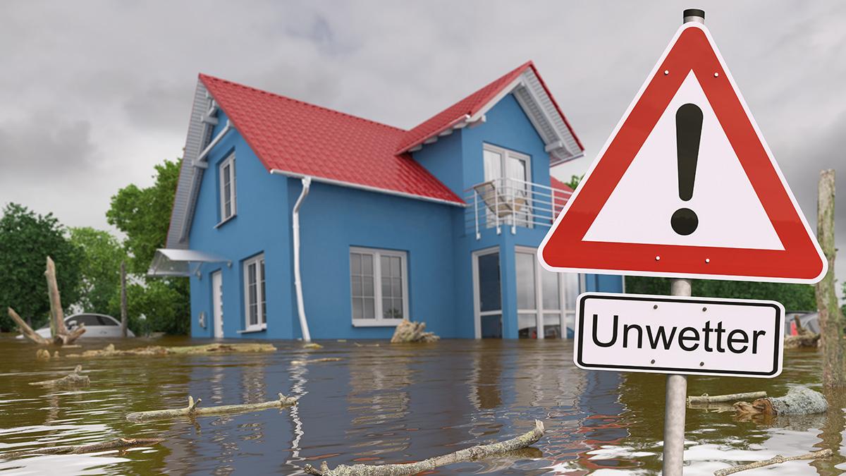 Hochwasser vor blautem Haus und ein Achtung-Verkehrsschild mit Zusatz "Unwetter"