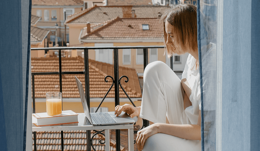 Frau sitzt mit Laptop an einem Tisch auf einem Balkon
