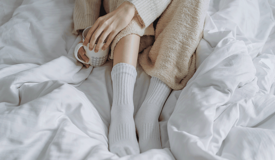 Person sitzt in Kuschelsocken und Hausanzug mit Heißgetränk auf Bett 