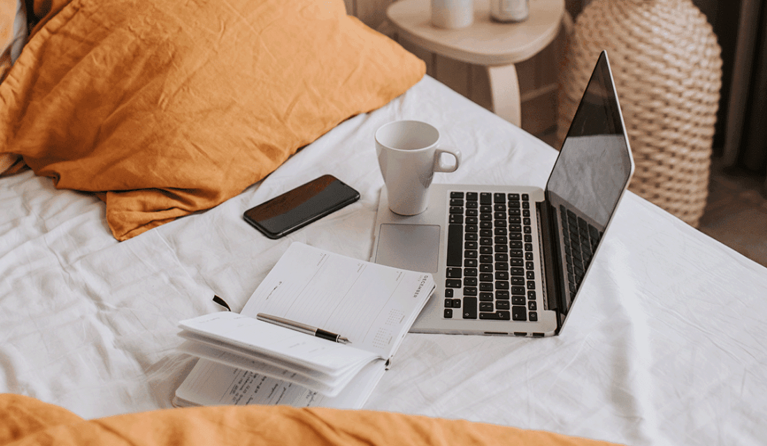 Auf einem Bett liegen Laptop, Kalender, Tasse und Handy