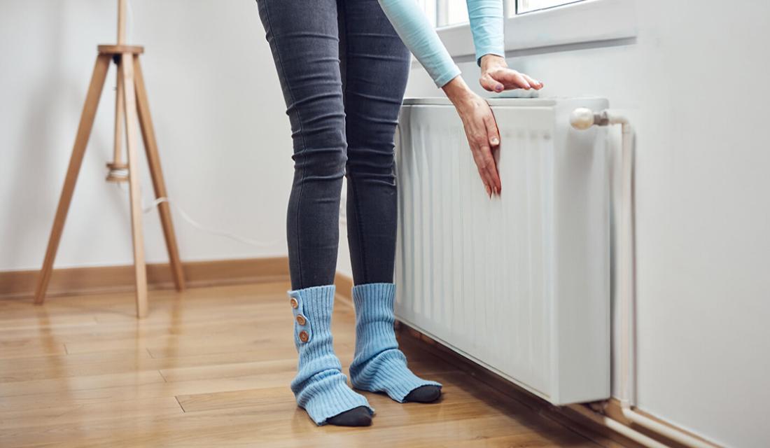 Frau steht vor einem Heizkörper und fühlt daran. An den Füßen hat sie Stulpen an.