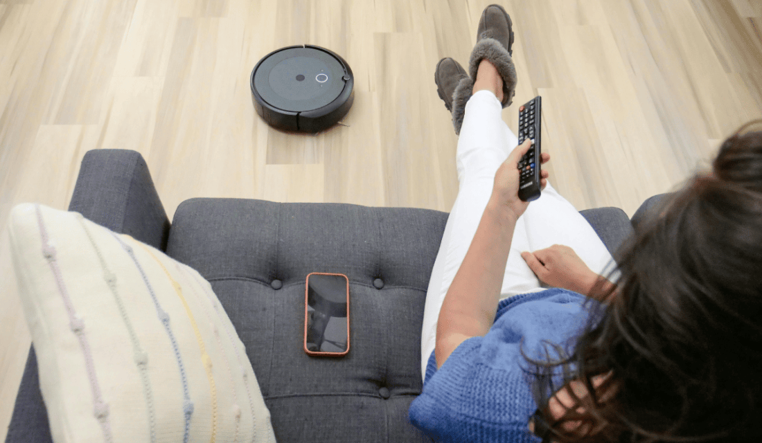 Blick von leicht oben über die Schulter einer Person, die auf einem Sofa sitzt und eine Fernbedienung nutzt. Neben ihr liegen ein Smartphone und ein Kissen, auf dem Boden befindet sich ein Staubsaugerroboter