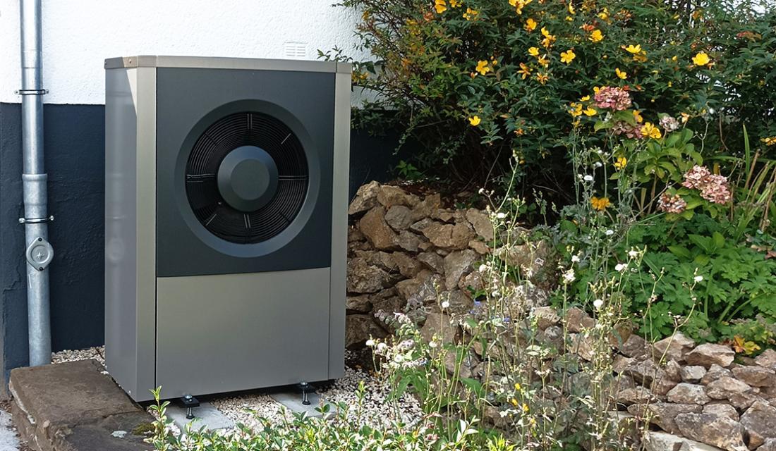 Graue Wärmepumpe vor einem Einfamilienhaus mit Blumen