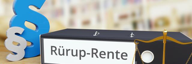 Ein Aktenordner mit der Aufschrift "Rürup-Rente" liegt auf einem Tisch, links daneben zwei Paragraph-Zeichen.