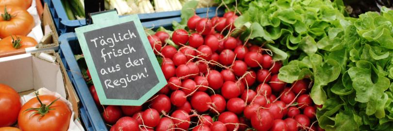 Auf einem Marktstand liegen Gemüse und Salate aus, ein Schild weist auf regionale Hersteller hin.