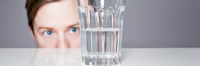 Ein Glas mit Wasser steht auf einer weißen glänzenden Platte, dahinter guckt ein Mann aufs Glas. Sein Gesicht ist oberhalb der Nasenspitze zu sehen.