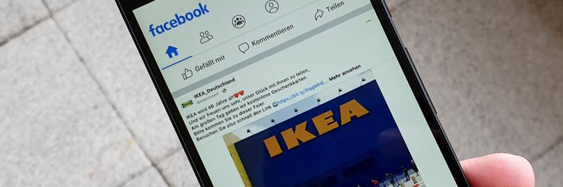 Hand hält Smartphone, Display zeigt einen Beitrag mit dem Foto eines Ikea-Gebäudes auf Facebook
