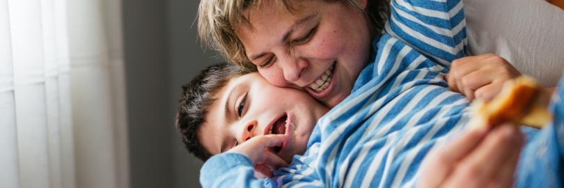 Eine Mutter liegt mit ihrem Sohn mit Behinderung auf einer hellen Decke; sie umarmt ihn liebevoll und kitzelt ihn, während beide herzhaft lachen und Freude ausstrahlen.