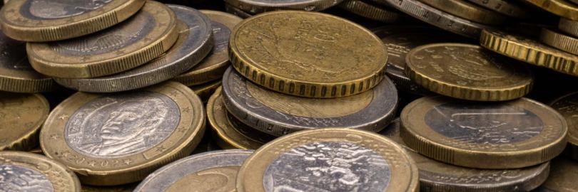 Eine Nahaufnahme zeigt einen großen, ungeordneten Haufen verschiedener Euro-Münzen, die übereinandergestapelt sind. Im Vordergrund sind deutlich 1-Euro- und 2-Euro-Münzen sowie goldfarbene Cent-Stücke (wahrscheinlich 50 Cent) zu erkennen. Die Münzen weisen Gebrauchsspuren und eine matte Textur auf, wobei das Licht die geprägten Ränder und Oberflächendetails hervorhebt.