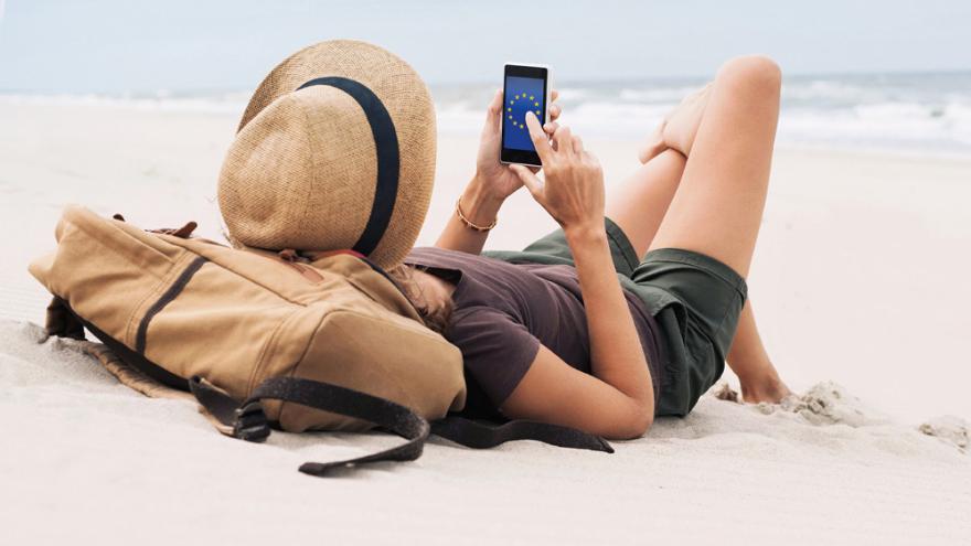 Eine Frau liegt im weißen Sand eines Strandes, Wellen unscharf im Hintergrund. In der Hand hält sie ein Smartphone mit der EU-Flagge auf dem Display.