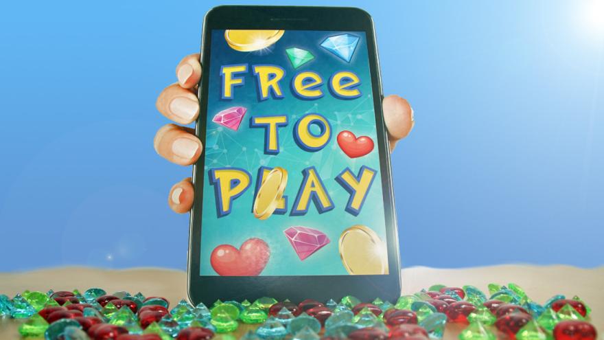 Eine Hand hält ein Smartphone mit der Aufschrift "FREE TO PLAY". Um die Schrift sind viele Münzen und Diamanten zu sehen, das Gerät steht auf einem Sandstrand mit Diamanten.