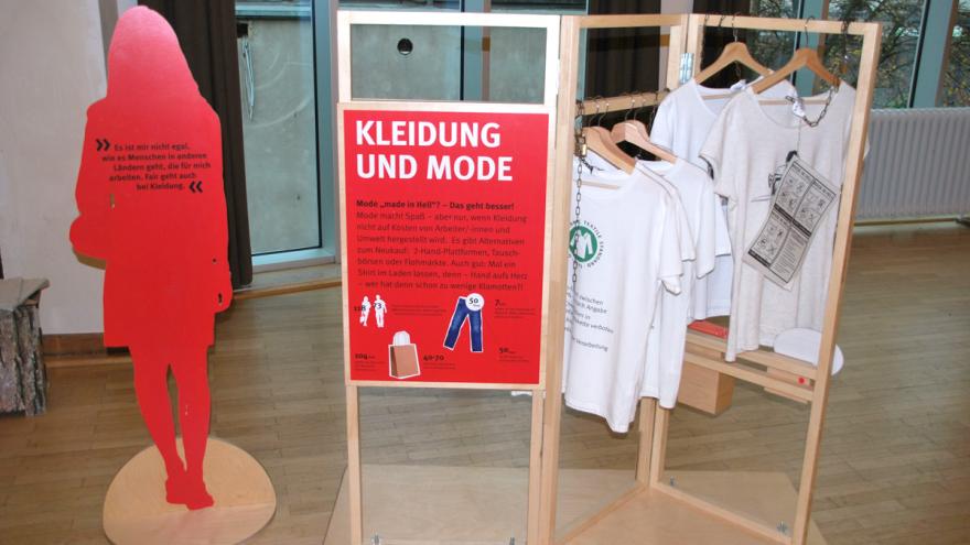 Umriss einer Frau als roter Holzaufsteller mit Text und Garderobe mit weißen T-Shirts