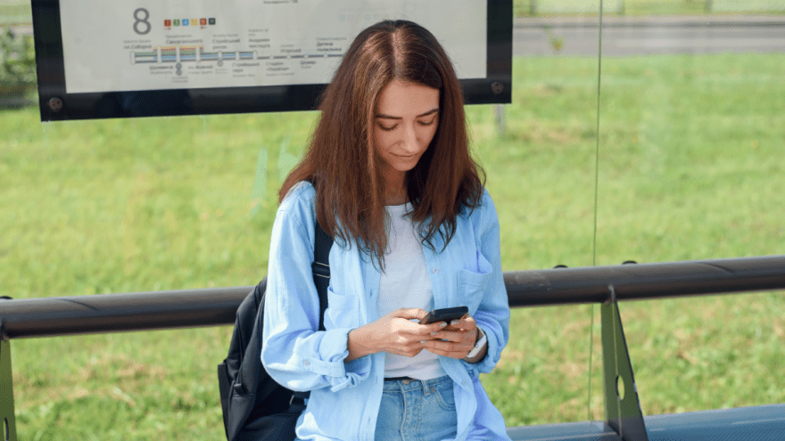 Eine junge Frau wartet an einer Bushaltestelle und schaut auf ihr Handy
