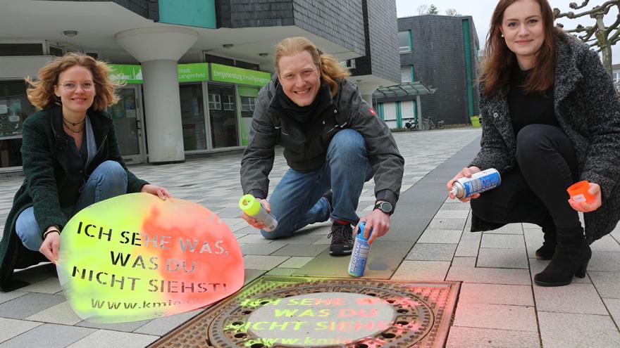 Drei Personen (zwei Frauen und ein Mann) mit Spraydosen an einem Kanaldeckel