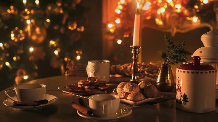 Gedeckter Tisch mit Kerze, Tassen, Plätzchen, im Hintergrund ein Tannenbaum mit leuchtenden Lichtern