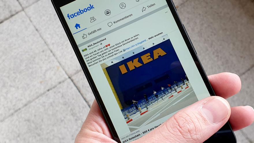 Hand hält Smartphone, Display zeigt einen Beitrag mit dem Foto eines Ikea-Gebäudes auf Facebook
