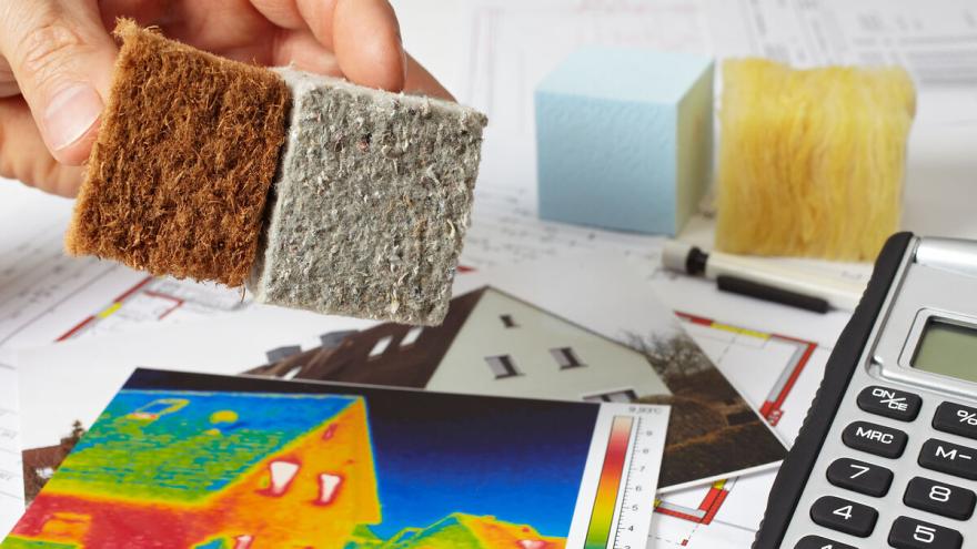 Mehrere Unterlagen wie eine Thermografie-Aufnahme eines Hauses liegen auf einer Oberfläche sowie ein Taschenrechner und Dämmstoffe. Eine Hand ragt ins Bild, welche zwei weitere Dämmstoffe hält.