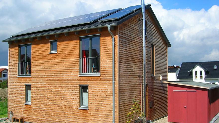 Kleines Einfamilienhaus mit zwei Etagen und Holzfassade, daneben ein kleiner Holzschuppen, auf dem Dach eine Solarthermie- und eine Photovoltaik-Anlage