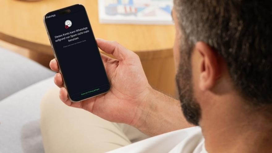 Ein Mann hält ein Smartphone in der Hand, auf dessen Disyplay in weißer Schrift auf schwarzem Hintergrund steht: "Dieses Konto kann WhatsApp aufgrund von Spam nicht mehr benutzen. Chats sind noch auf diesem Gerät."