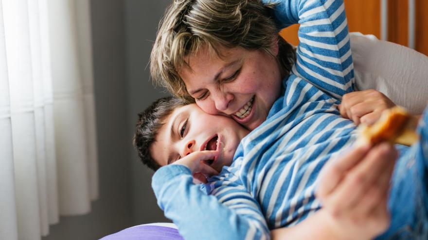 Eine Mutter liegt mit ihrem Sohn mit Behinderung auf einer hellen Decke; sie umarmt ihn liebevoll und kitzelt ihn, während beide herzhaft lachen und Freude ausstrahlen.
