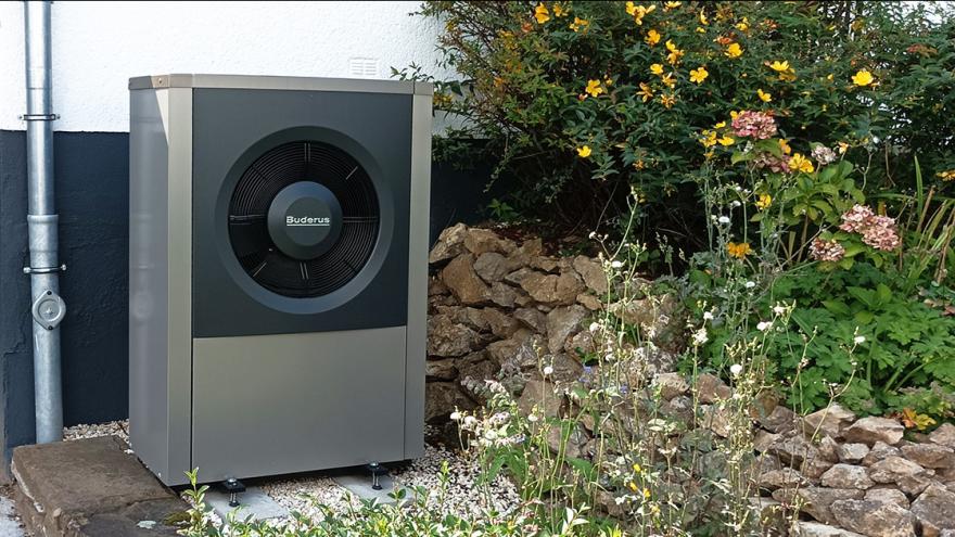 Graue Wärmepumpe vor einem Einfamilienhaus mit Blumen