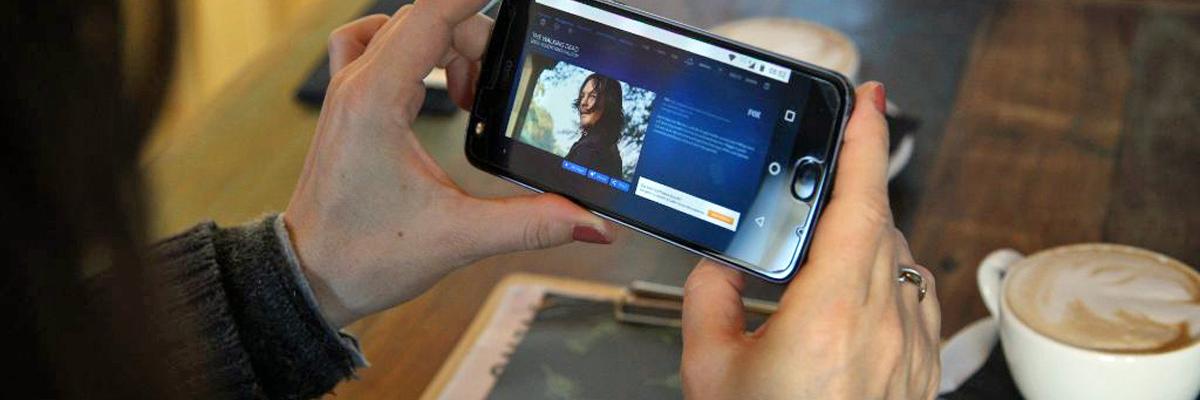 Eine Frau hält in einem Café ein Smartphone in der Hand, auf dem ein Video-Streaming-Dienst zu sehen ist.