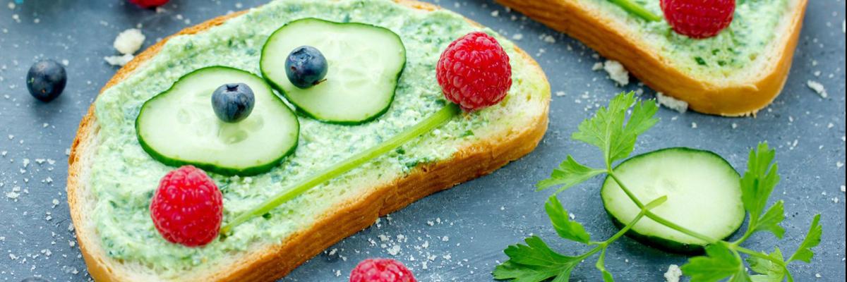 Gurkenscheiben, Petersilienstängel und Himbeeren bilden Gesichter auf belegten Brotscheiben.