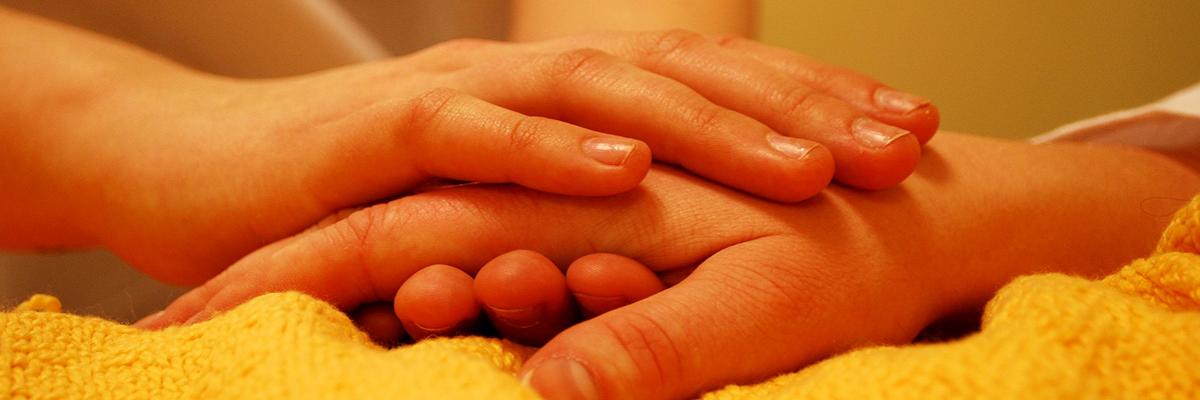 Hände einer pflegenden Person halten die Hand einer im Bett liegenden Person
