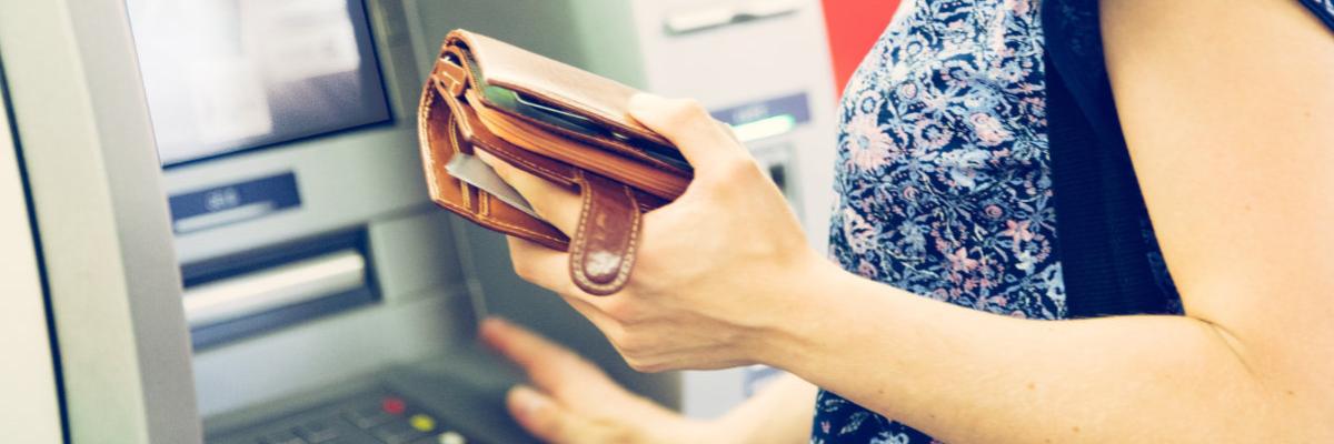 Eine Frau hält eine Geldbörse in der Hand und steht vor einem Geldautomaten
