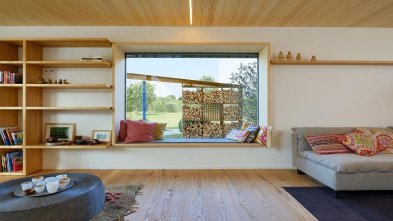 Blick aus Wohnraum durch ein großes Sitzfenster in den Garten auf aufgeschichtetes Holz