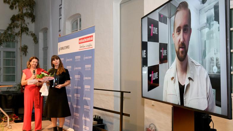 Eva Eisemann und Nina Kannenberg stehen auf der rechten Bildseite etwas im Hintergrund. Links ist ein großer Bildschirm mit dem Gesicht von Philip Strunk zu sehen.