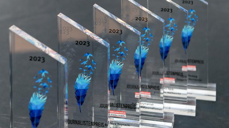 Sechs Trophäen aus Acrylglas mit der Zahl 2023 über einer blauen Feder, Namen der Preisträger:innen und Logo der Verbraucherzentale Nordrhein-Westfalen
