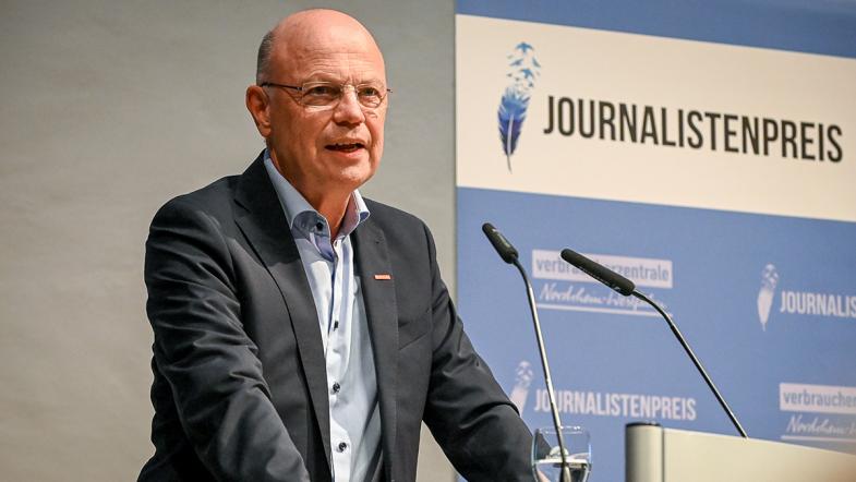 Wolfgang Schuldzinski steht an einem Rednerpult, hinter ihm ein Banner mit der Aufschrift Journalistenpreis