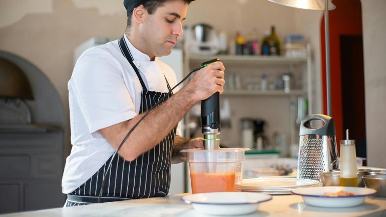 Mann mit Kochschürze hält einen Pürierstab in einen Behälter mit orangefarbener Flüssigkeit