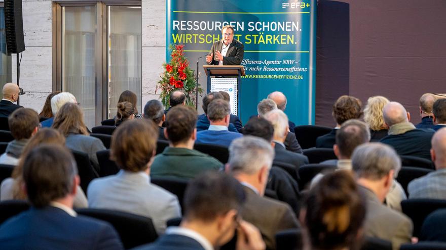 Ein Mann steht in der Ferne am Rednerpult, im Vordergrund sitzen zahlreiche Gäste.