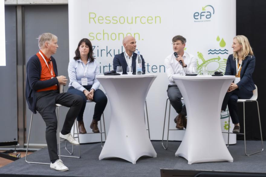 5 Leute an Tischen bei Diskussion auf demPosium