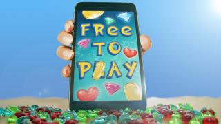Eine Hand hält ein Smartphone mit der Aufschrift "FREE TO PLAY". Um die Schrift sind viele Münzen und Diamanten zu sehen, das Gerät steht auf einem Sandstrand mit Diamanten.