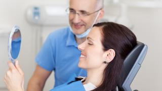 Frau sitzt beim Zahnarzt und schaut sich im Spiegel zufrieden ihre Zähne an.