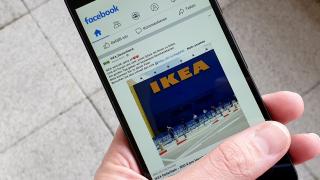 Hand hält Smartphone, Display zeigt einen Beitrag mit dem Foto eines Ikea-Gebäudes auf Facebook