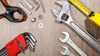 Zange, Dübel, Schrauben und Muttern, Maßband, Schraubenschlüssel und Imbusschlüssel liegen auf einer Tischplatte.