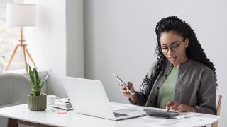 Foto einer Frau am Schreibtisch, die in einer Hand ein Handy hält und mit der anderen einen Taschenrechner bedient. Vor ihr steht ein geöffneter Laptop.