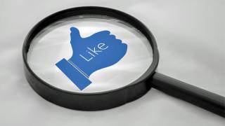 Eine Lupe liegt auf einer symbolisierten blauen Hand mit aufzeigendem Daumen mit der Aufschrift "Like"