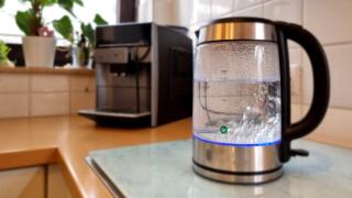 Zu kochen beginnendes Wasser in einem gläsernen Wasserkocher auf der Arbeitsplatte einer Küche, dahinter unscharf ein Kaffeevollautomat.