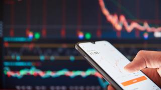 Nahaufnahme eines Smartphones, das in einer Hand gehalten wird und einen roten, absteigenden Graphen auf dem Bildschirm anzeigt. Im Hintergrund ist ein größerer Bildschirm mit einem ähnlichen Graphen zu sehen, der ebenfalls einen Abwärtstrend zeigt. 