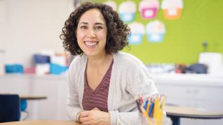 Eine Frau sitzt in einem Klassenzimmer an einem Tisch, stützt sich mit gefalteten Händen auf ihn und lächelt in die Kamera