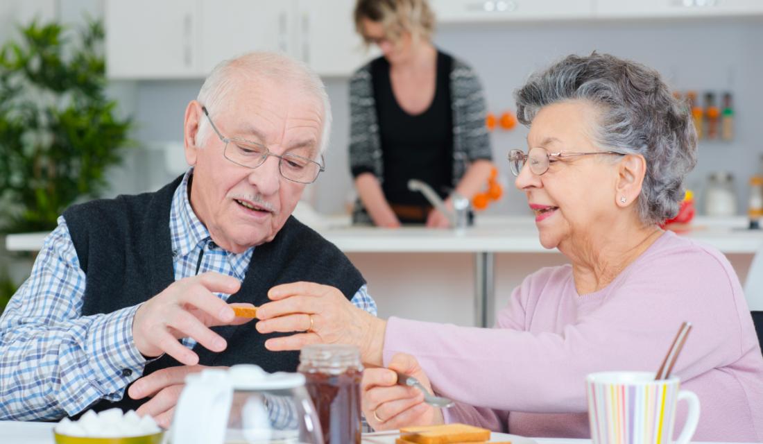 Ein älteres Paar sitzt am Tisch und frühstückt, während im Hintergrund eine junge Frau in der Küche arbeitet.