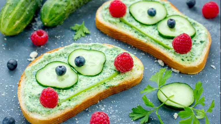 Gurkenscheiben, Petersilienstängel und Himbeeren bilden Gesichter auf belegten Brotscheiben.