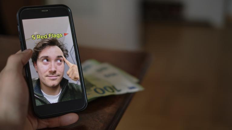 Eine Hand hält ein Smartphone im Hochformat vor einem unscharfen Hintergrund, auf dem Geldscheine (ein 100-Euro-Schein ist erkennbar) auf einem Holztisch liegen.  Auf dem Display des Smartphones ist ein junger Mann in einer Video-App zu sehen. Er blickt nachdenklich nach oben und tippt sich mit dem Zeigefinger an die Schläfe. Über seinem Kopf steht der gelbe Text „5 Red Flags“, gefolgt von einem kleinen roten Flaggen-Emoji.